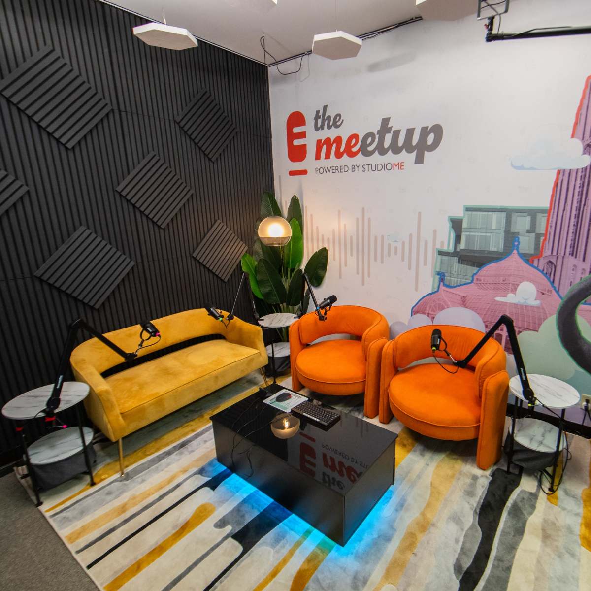 A podcast studio featuring two orange chairs and a yellow sofa arranged around a black coffee table, with a decorative wall mural and soundproofing panels in the background.