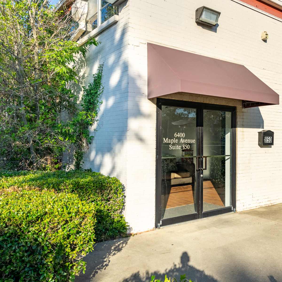 The entrance features a glass door with a brown awning, flanked by manicured shrubs, leading into an office suite.