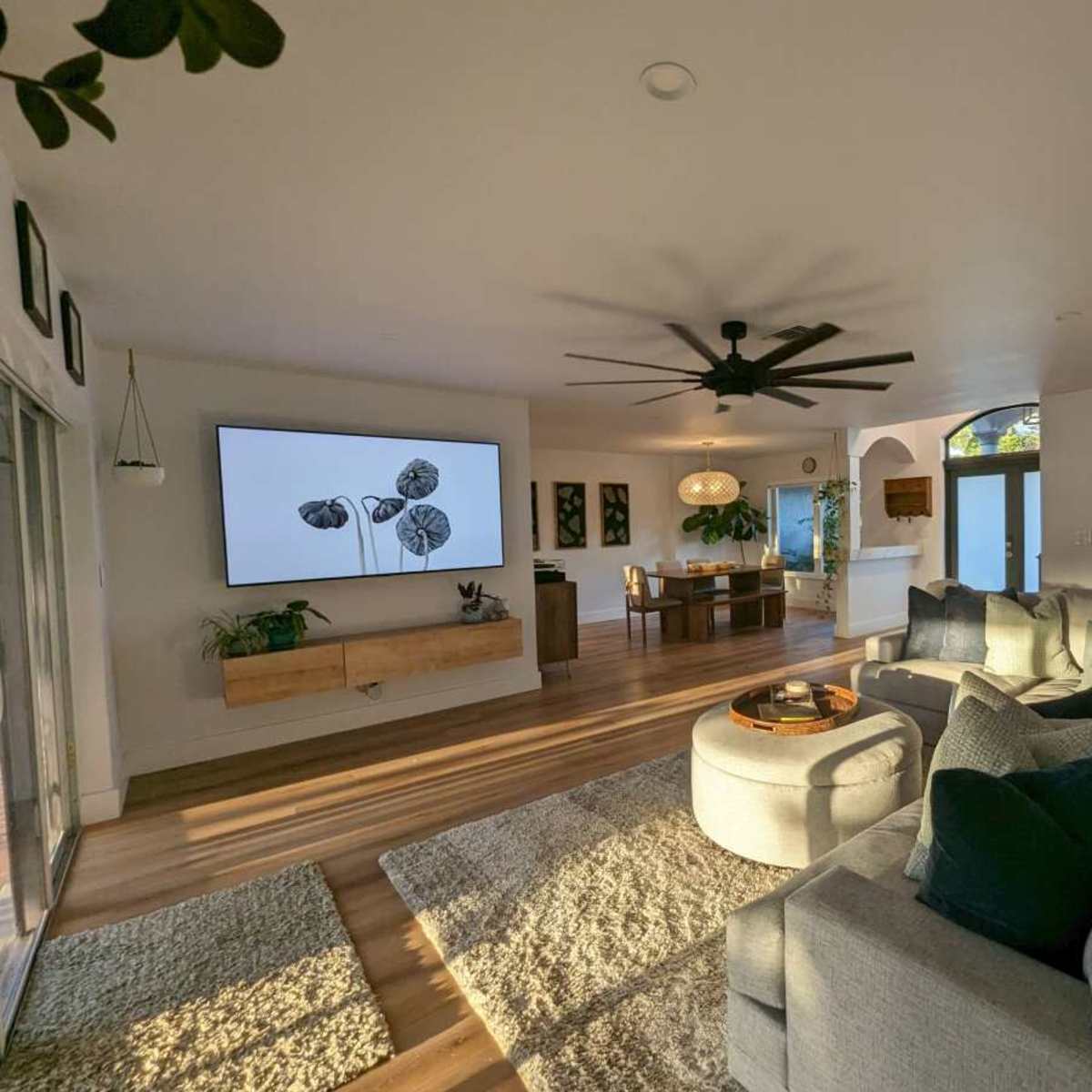 The living room features a large sectional sofa, a circular coffee table, a wall-mounted television displaying artwork, and a dining area in the background.