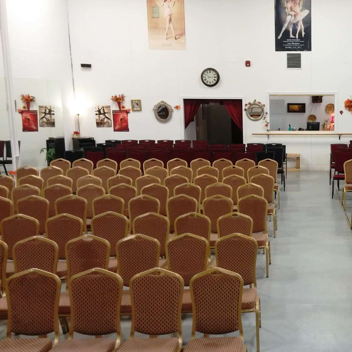 A large room with rows of empty, gold-framed chairs facing a stage area, decorated with various artworks and posters on the walls.