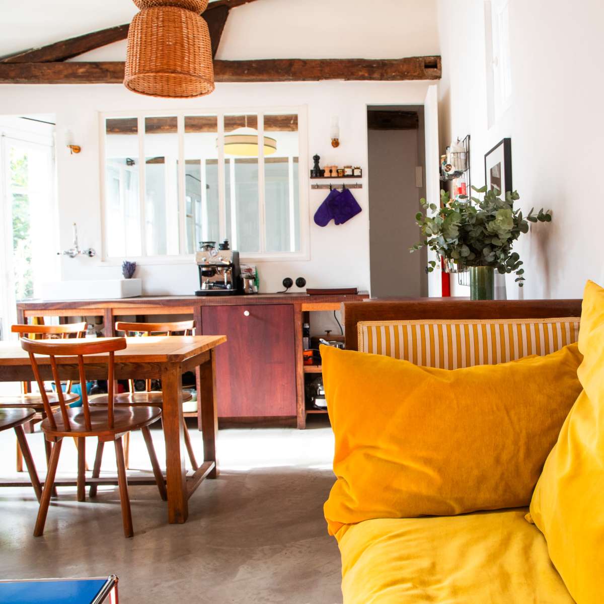 A bright, open living space with a yellow couch, a wooden dining table, and a kitchen area in the background.