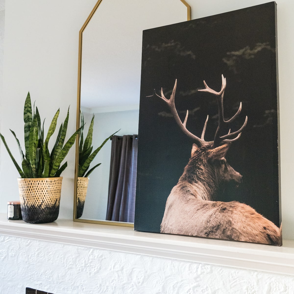 A mounted deer canvas is displayed above a fireplace, alongside a gold-framed mirror and potted plants.