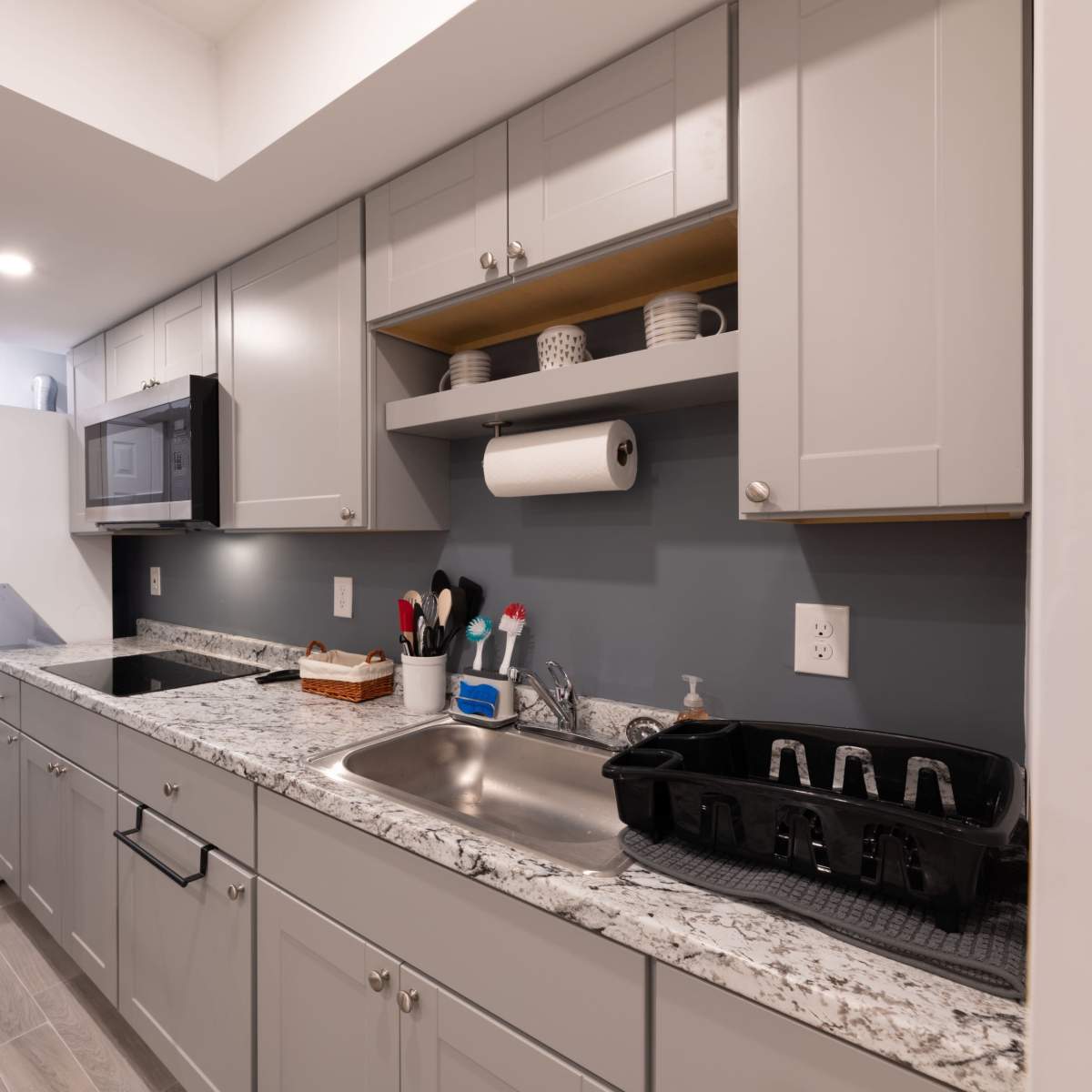A modern kitchen with gray cabinets, a granite countertop, a sink, and appliances including a microwave and a laundry area.