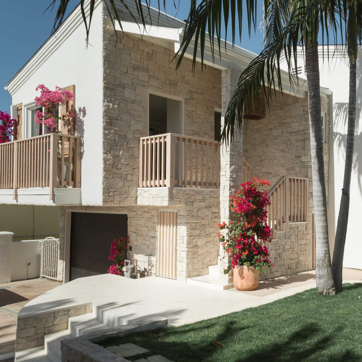 A modern two-story house with stone and white walls, featuring a balcony, palm trees, and vibrant flowers in large pots.