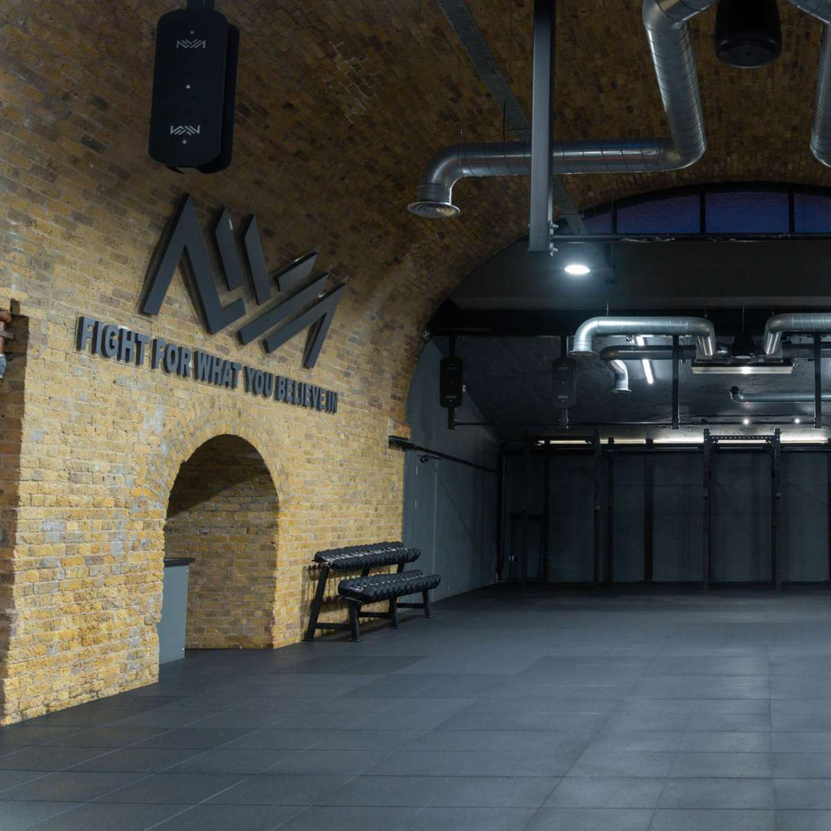 A spacious gym interior with exposed brick walls, high ceilings, and various workout equipment arranged in a clean, open layout.