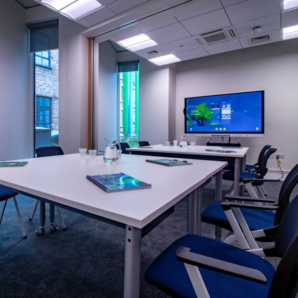 A modern conference room features a large table surrounded by chairs, with a television screen displaying content and sunlight streaming through windows.