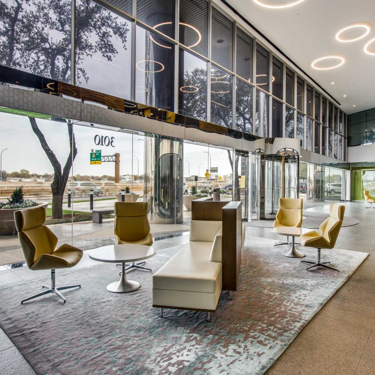 A modern lobby area featuring sleek chairs and a round table on a rug, with large glass windows allowing natural light and a view of the outside.