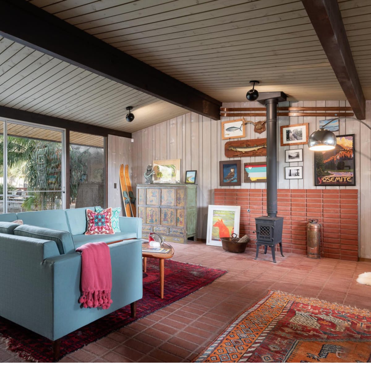 A cozy living room featuring a light blue sofa, a wood-burning stove, and various framed artworks on the walls, with large glass windows offering a view of a garden.