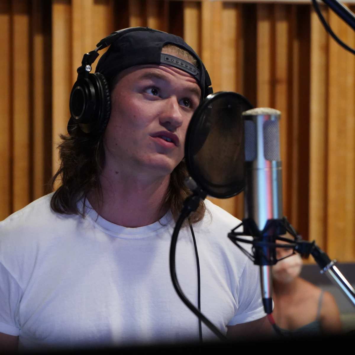 A young man with long hair wearing a white t-shirt and headphones speaks into a microphone in a recording studio.