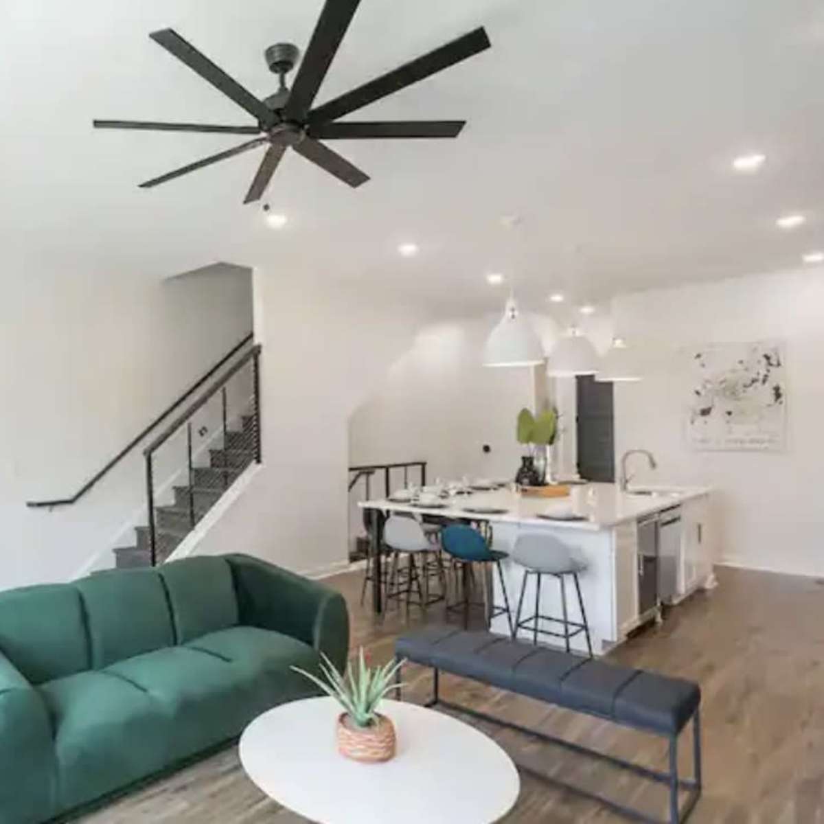 A modern living space with a green sofa, a round coffee table, a kitchen area featuring bar stools, and a staircase in the background.