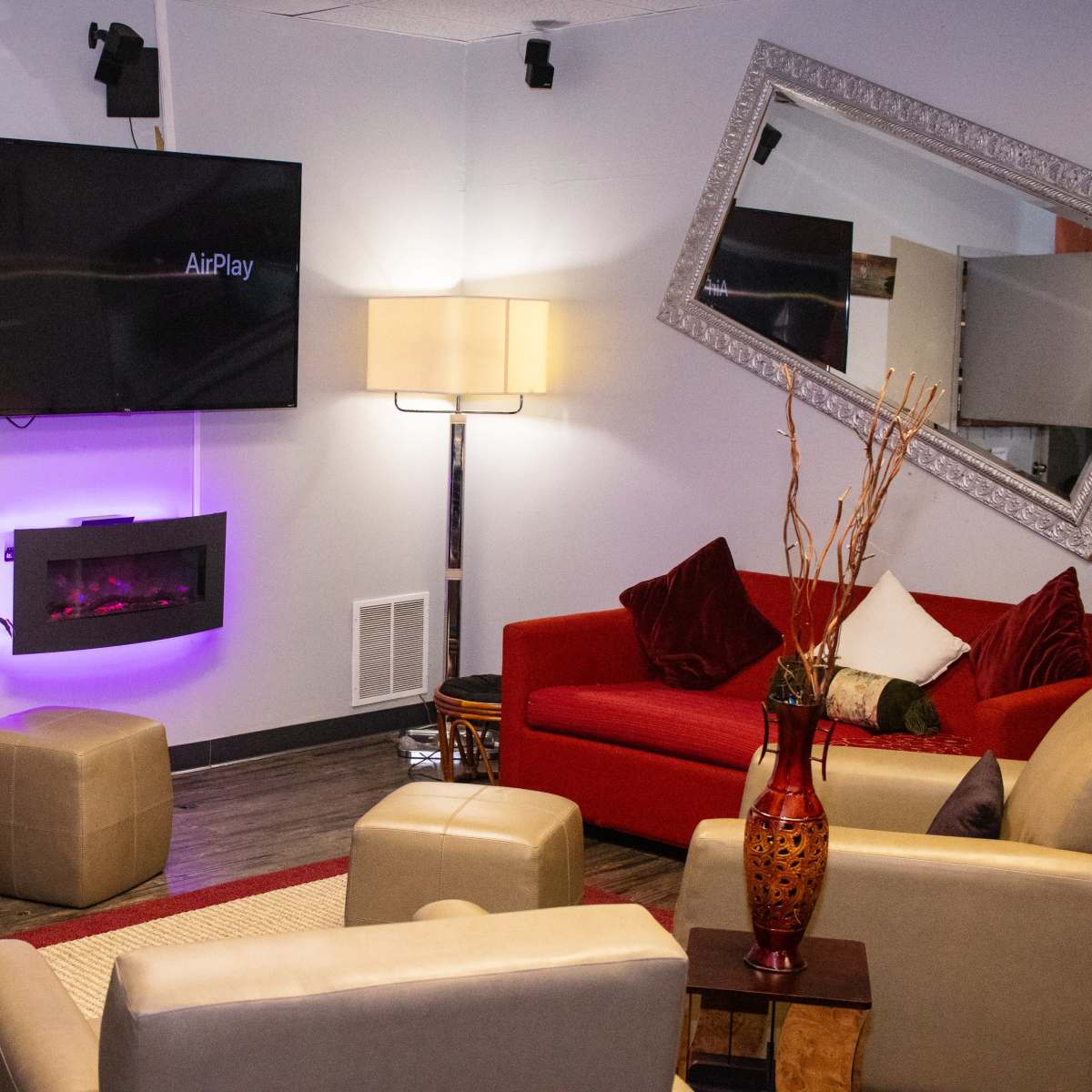 A cozy room features a TV mounted on the wall, a modern electric fireplace, a lamp, and various seating options including red and cream-colored chairs and ottomans.