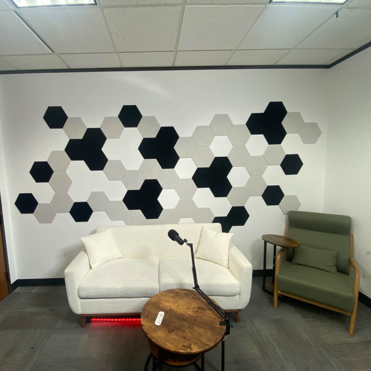 A small room with a white couch, a wooden side table, a microphone on a stand, and a wall adorned with hexagon-shaped acoustic panels in black and gray.