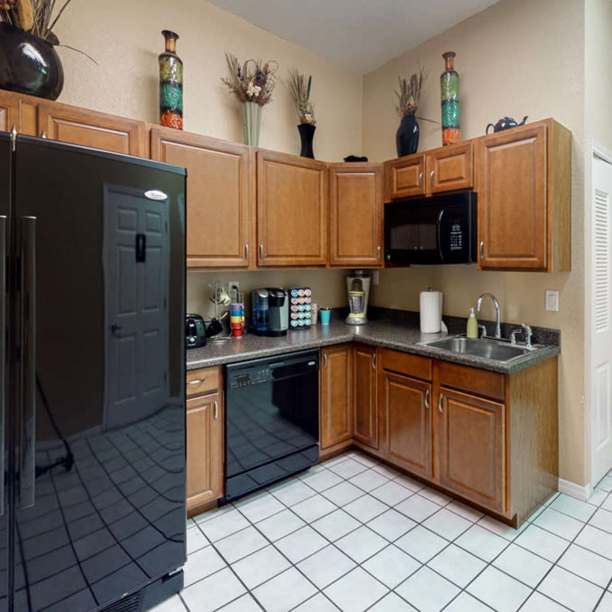 A kitchen equipped with wooden cabinets, a black refrigerator, a microwave, and a tiled floor, adjacent to a door leading outside.