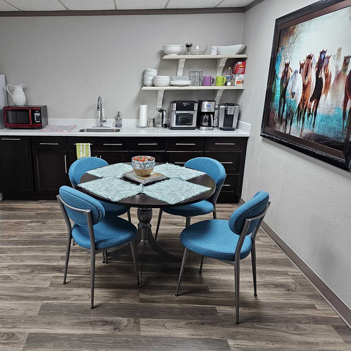 A small kitchen area with a round table and four blue chairs, along with a refrigerator, microwave, sink, and coffee maker.