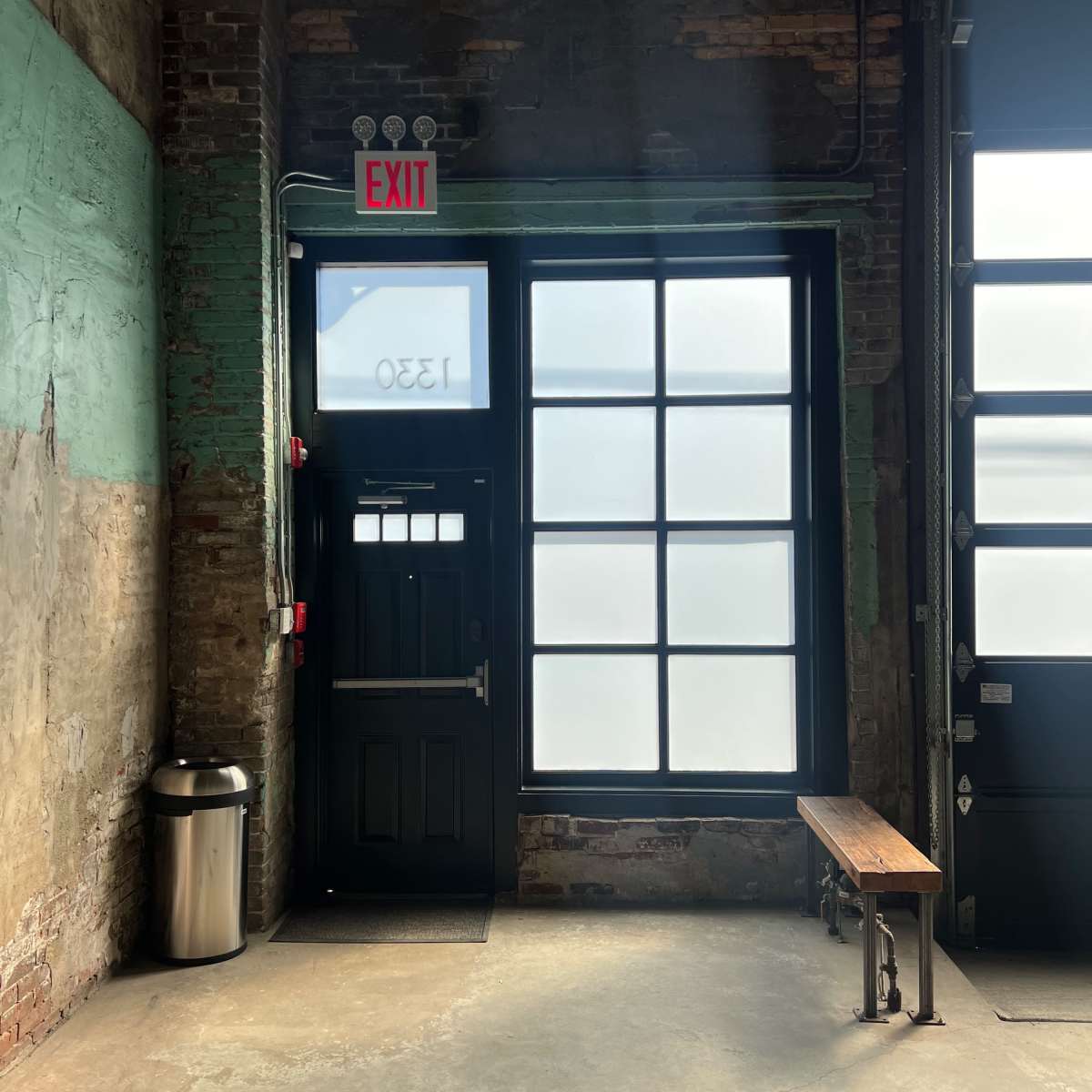 A room with exposed brick walls, featuring a large window and a black door with an exit sign, alongside a wooden bench and a trash can.