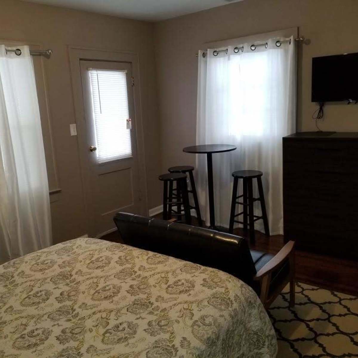 A small bedroom with a bed, a set of stools around a tall table, a dresser, and windows with white curtains.