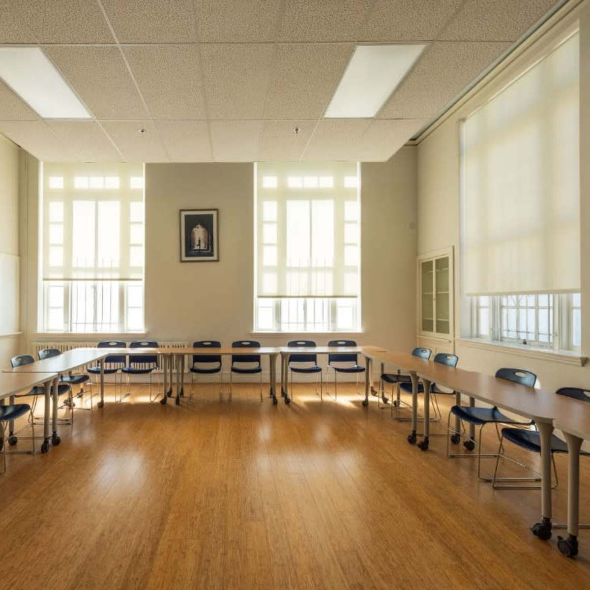 A spacious classroom is arranged with tables and chairs in a circular formation, illuminated by large windows.