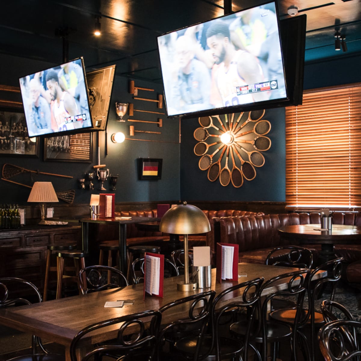 A cozy bar interior featuring leather seating, wooden tables, and two televisions displaying a sports game on the walls.