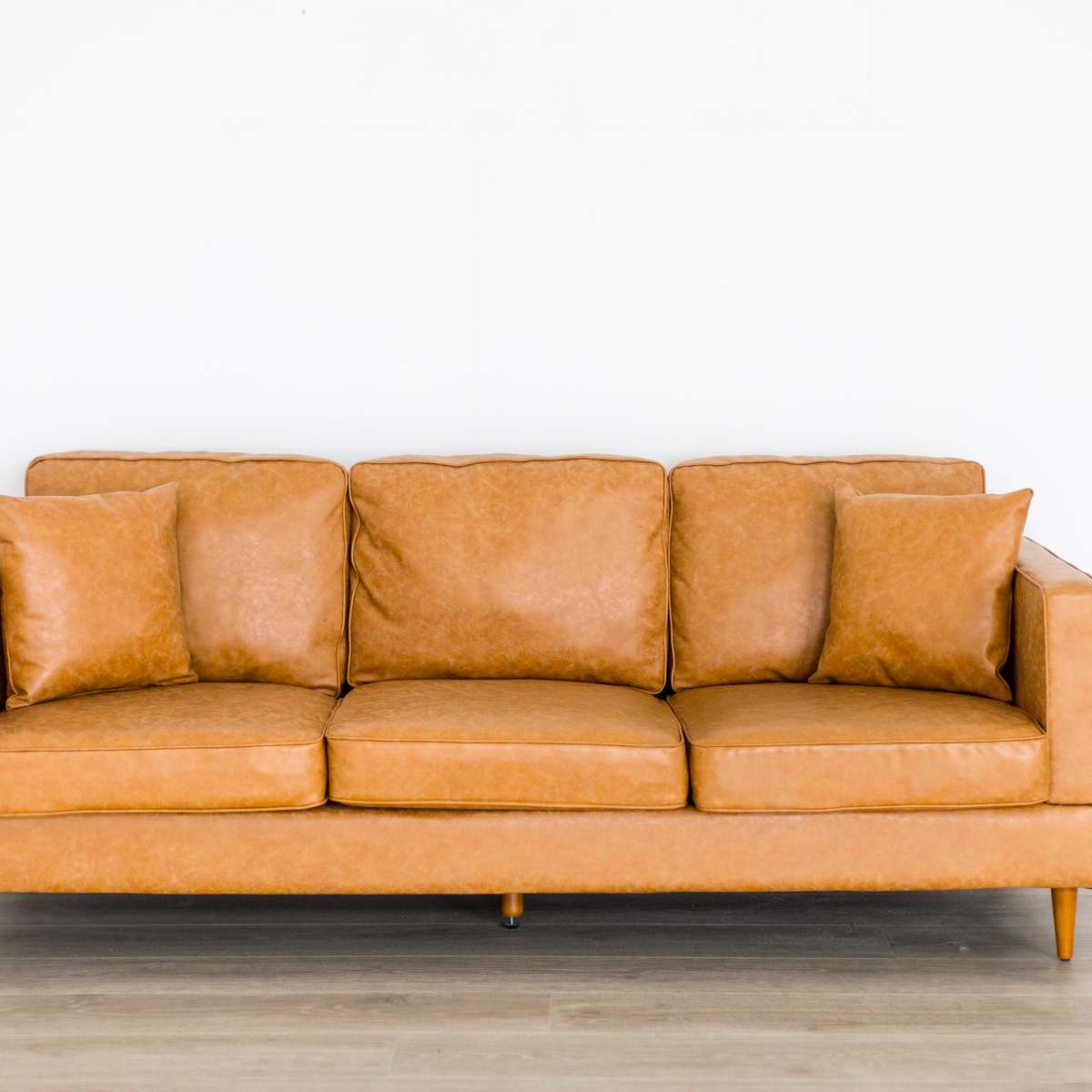 A tan leather sofa with three seat cushions and two matching pillows is positioned against a white wall.