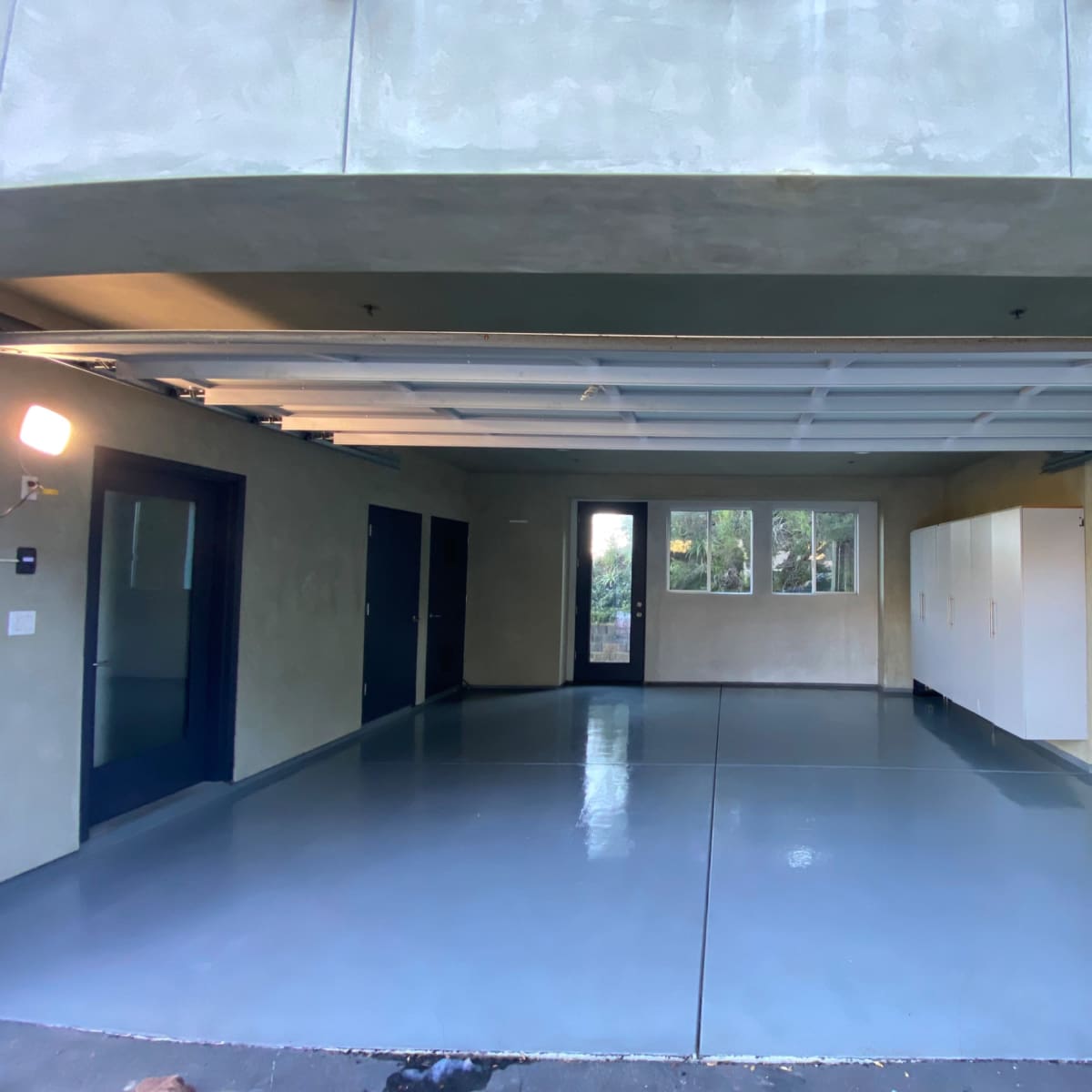 A spacious, empty garage with a grey floor and walls, featuring large windows and white storage cabinets.