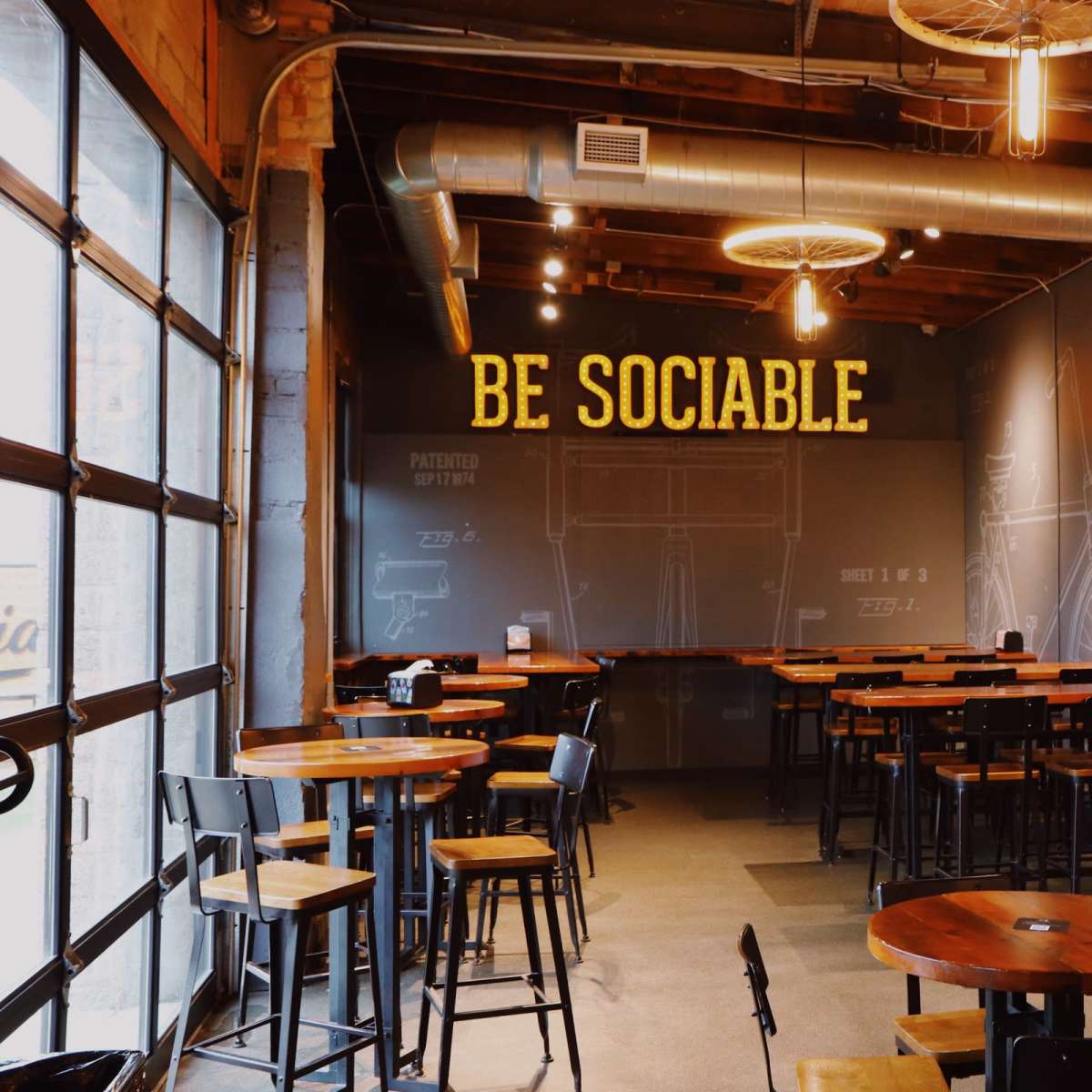 A modern café interior with wooden tables, high chairs, and a large wall sign that reads "BE SOCIABLE."