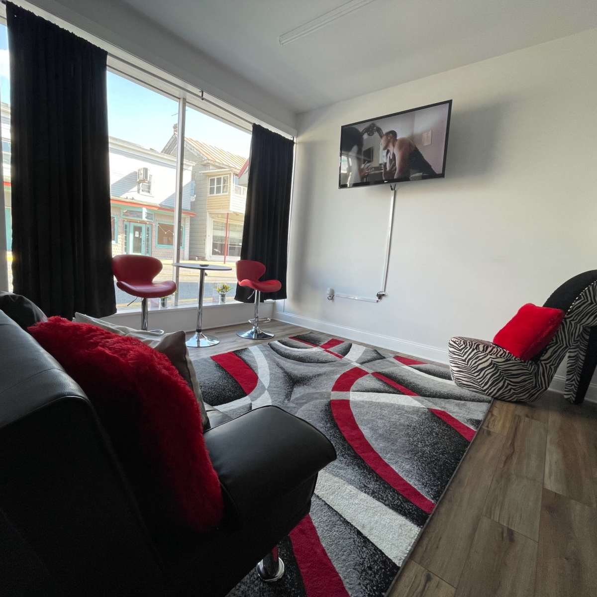 A modern living room with a large window, a black sofa, red and white decor, and two bar stools facing a wall-mounted television.