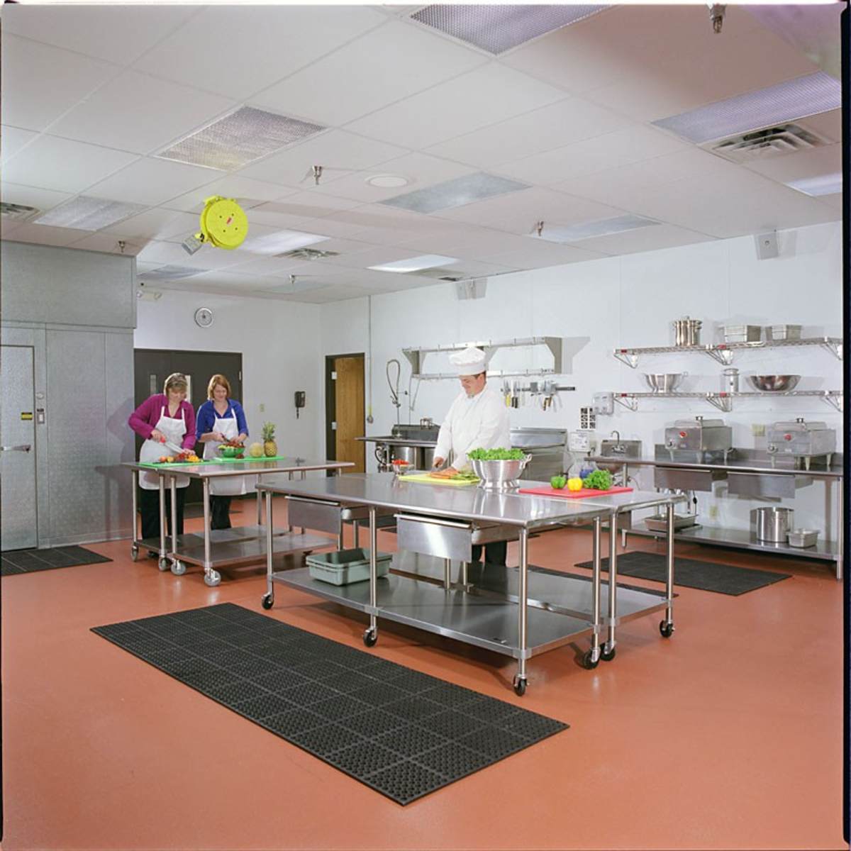 A clean, organized kitchen with stainless steel work tables, a chef in a white uniform, and two women preparing ingredients nearby.