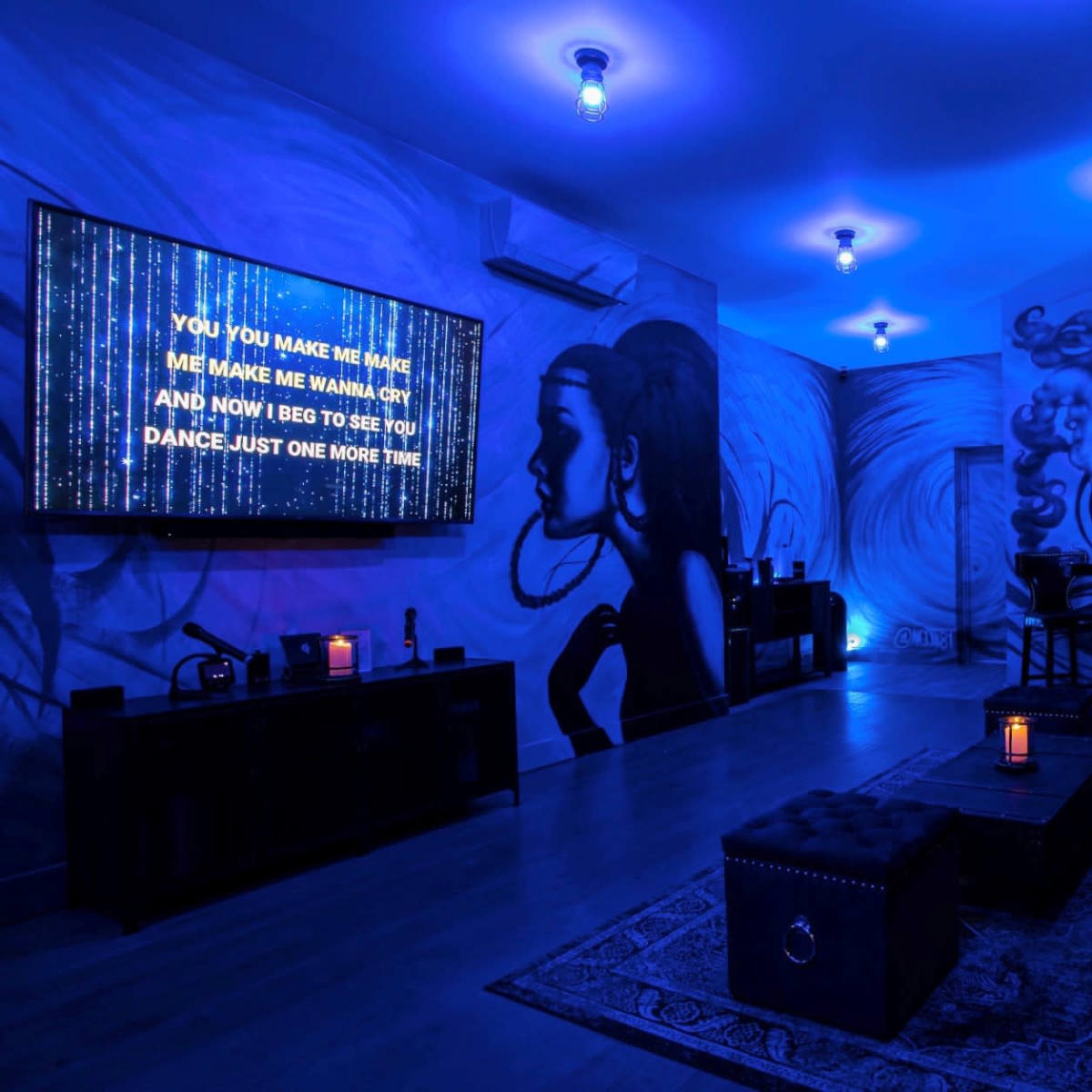 The room features blue lighting, a large television displaying lyrics, and walls adorned with artistic murals of women.