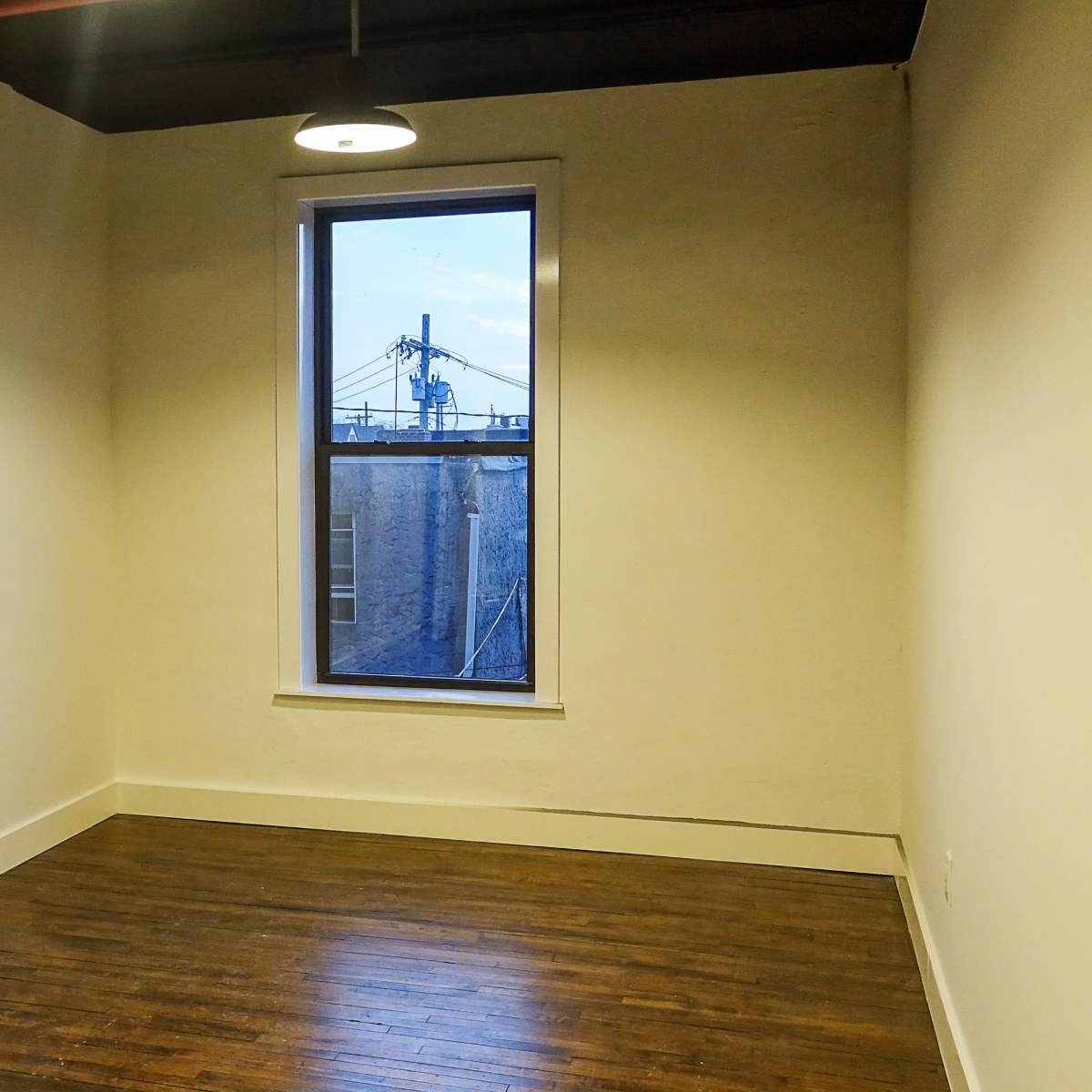 An empty room with dark wood flooring, a single window, and a light fixture on the ceiling.