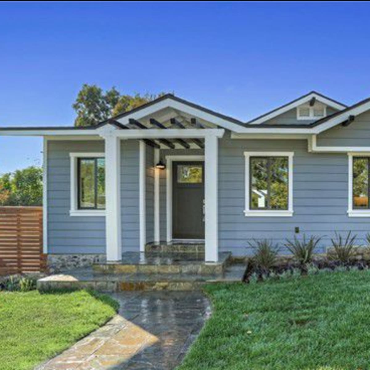 A single-story blue house with a stone pathway, a well-manicured lawn, and wooden fencing.