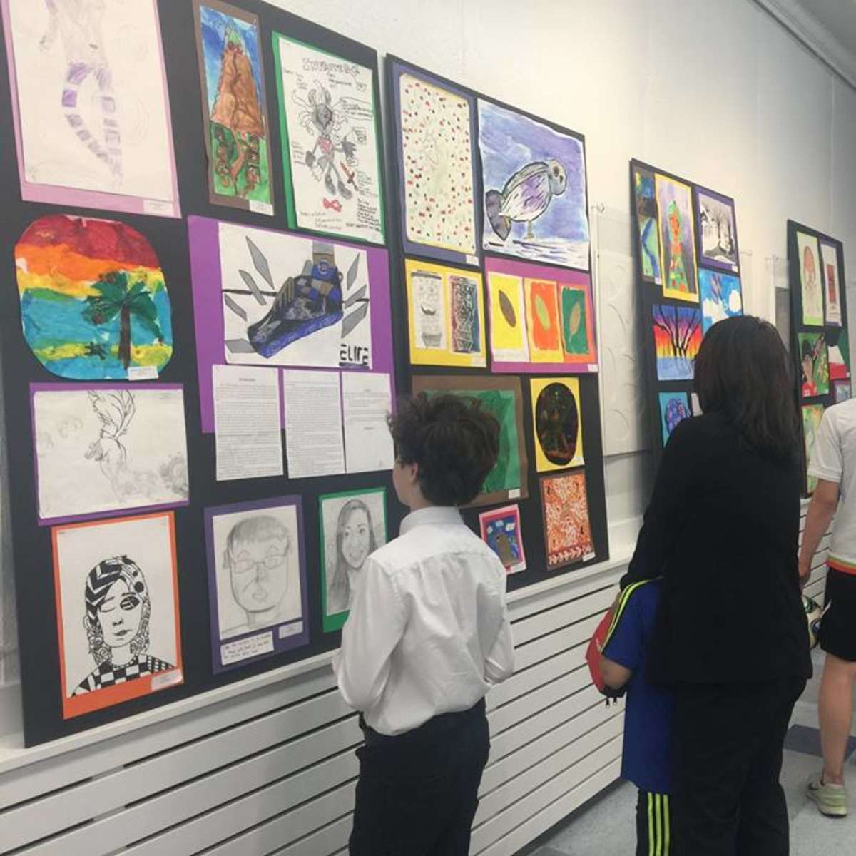 A group of people views artwork displayed on the walls of a school hallway during an exhibition.