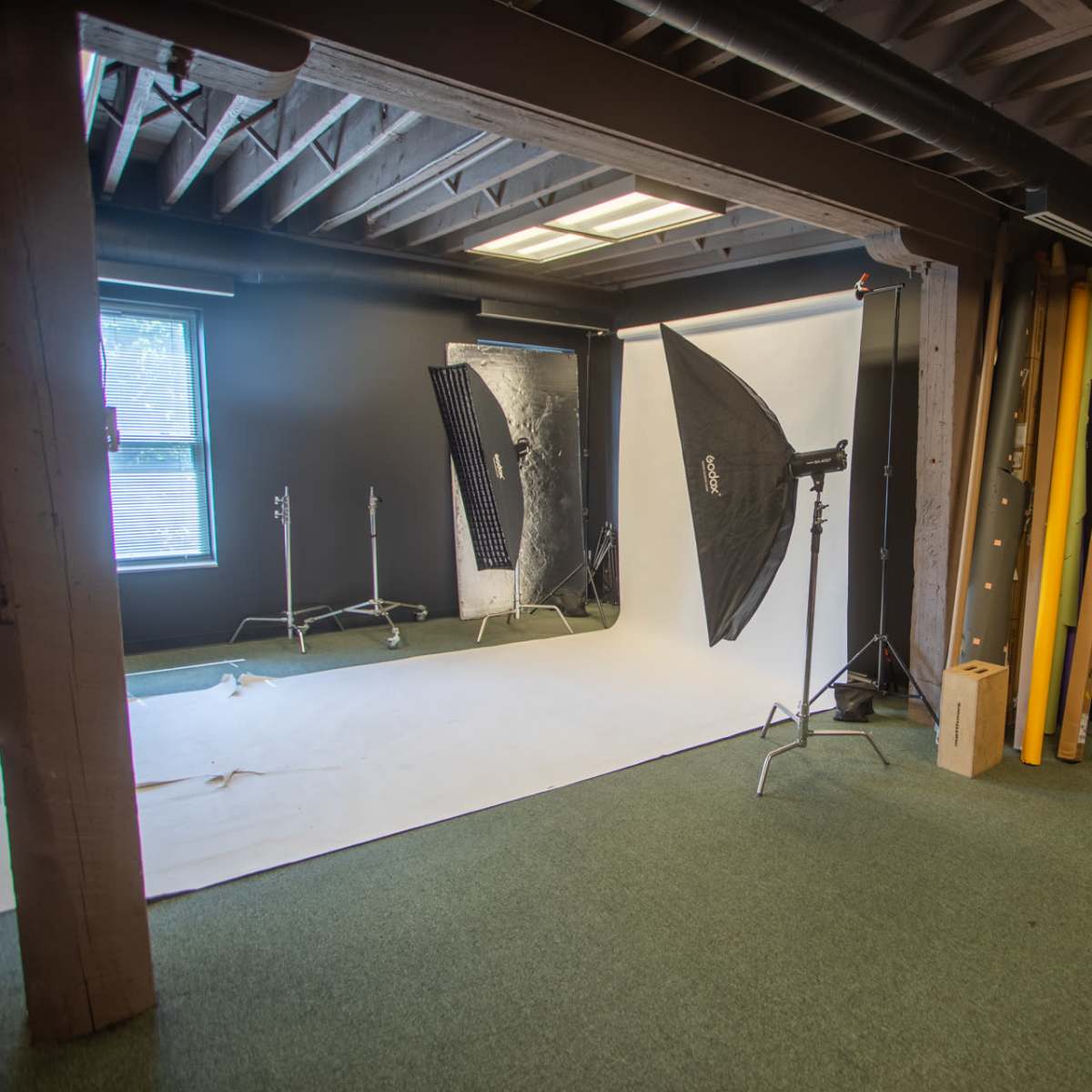 A photography studio featuring a white backdrop, two softbox lights on stands, and a dark carpeted area with a black couch and additional props in the corner.