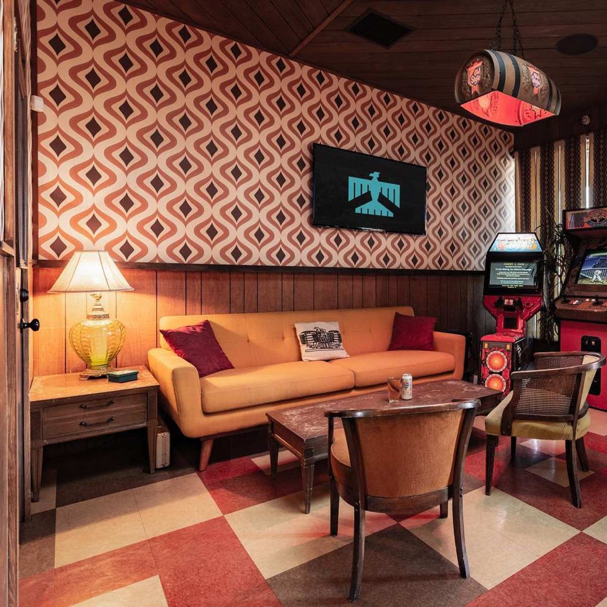 A cozy retro-themed lounge features a yellow sofa, wooden walls, and vintage arcade machines against a patterned wallpaper backdrop.