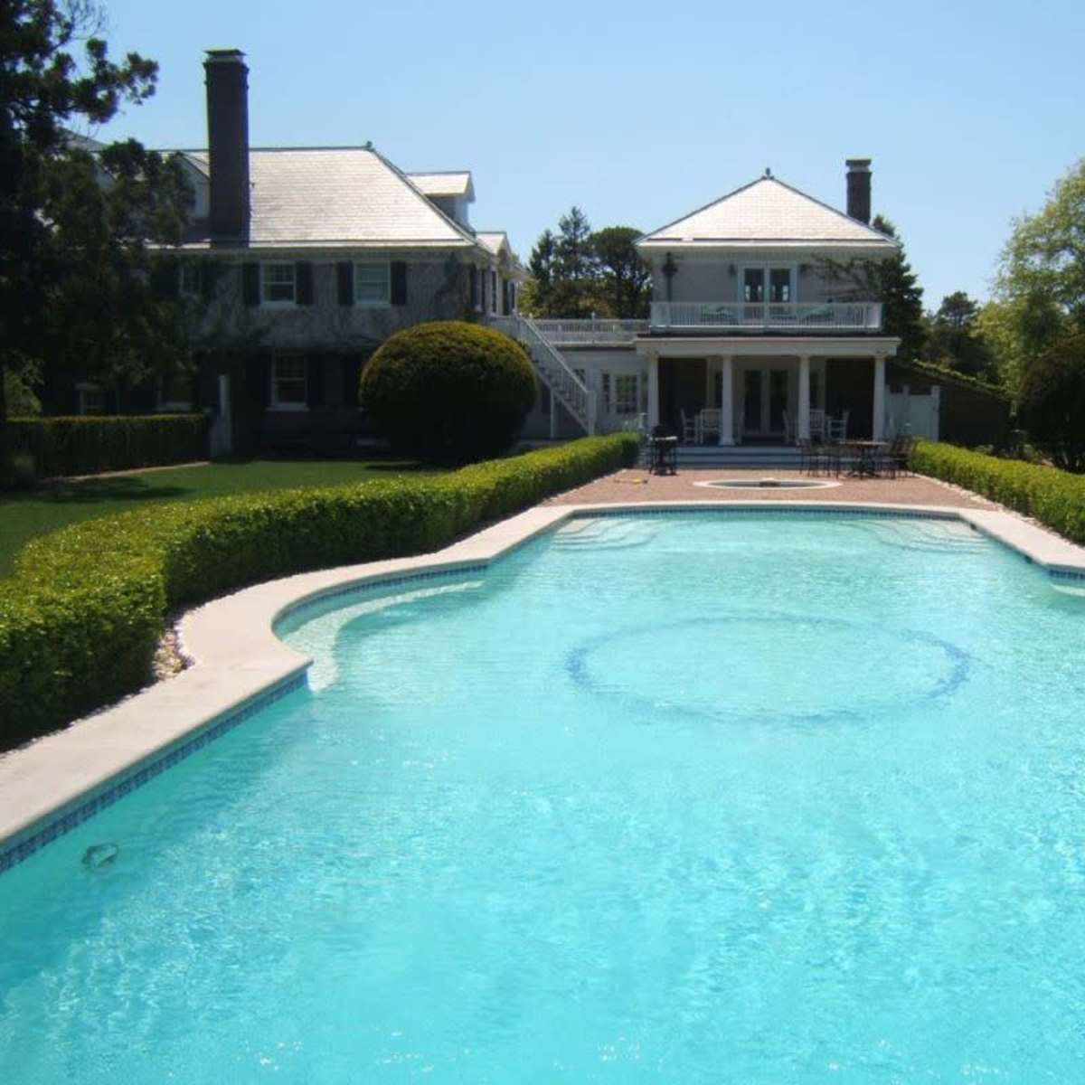 A clear blue swimming pool is situated in front of a large house, surrounded by well-manicured lawns and hedges.