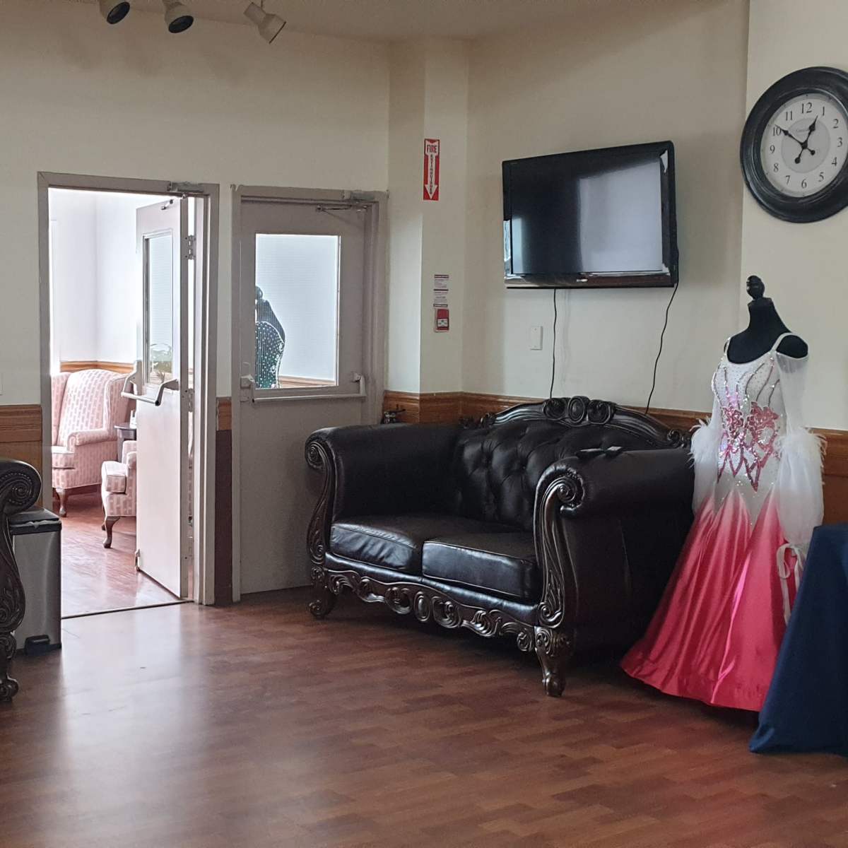 The scene features two black leather sofas, a doorway leading to another room, a wall clock, and a formal dress displayed on a mannequin.