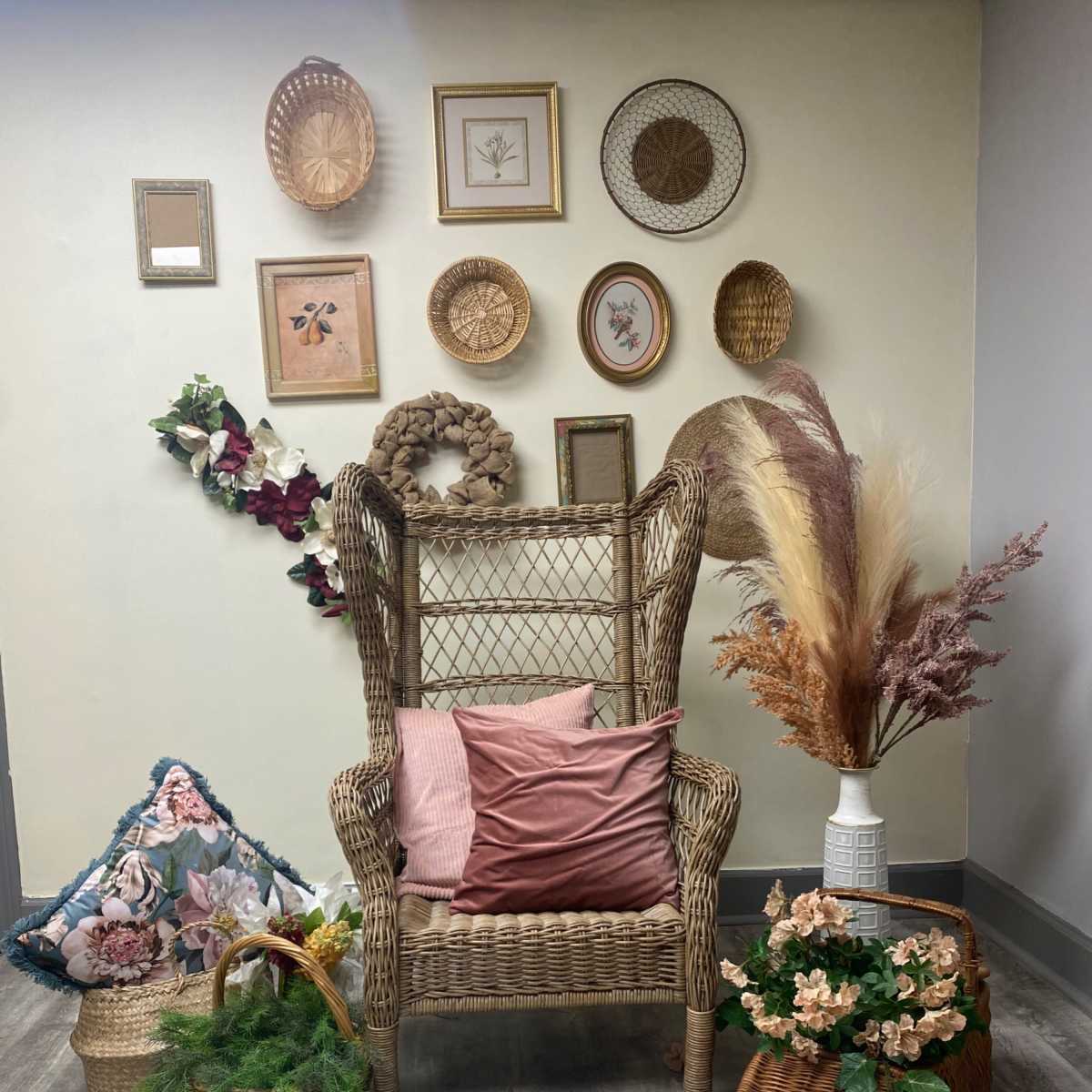 A wicker chair is surrounded by decorative baskets, floral arrangements, and framed pictures against a wall.