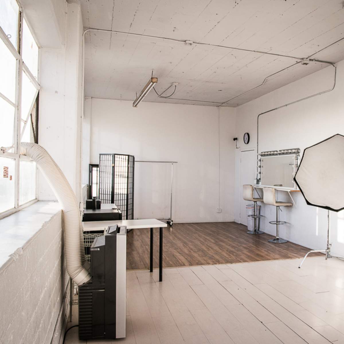 A photography studio with large windows, a light-colored floor, and various equipment including a backdrop, a table, and lighting modifiers.