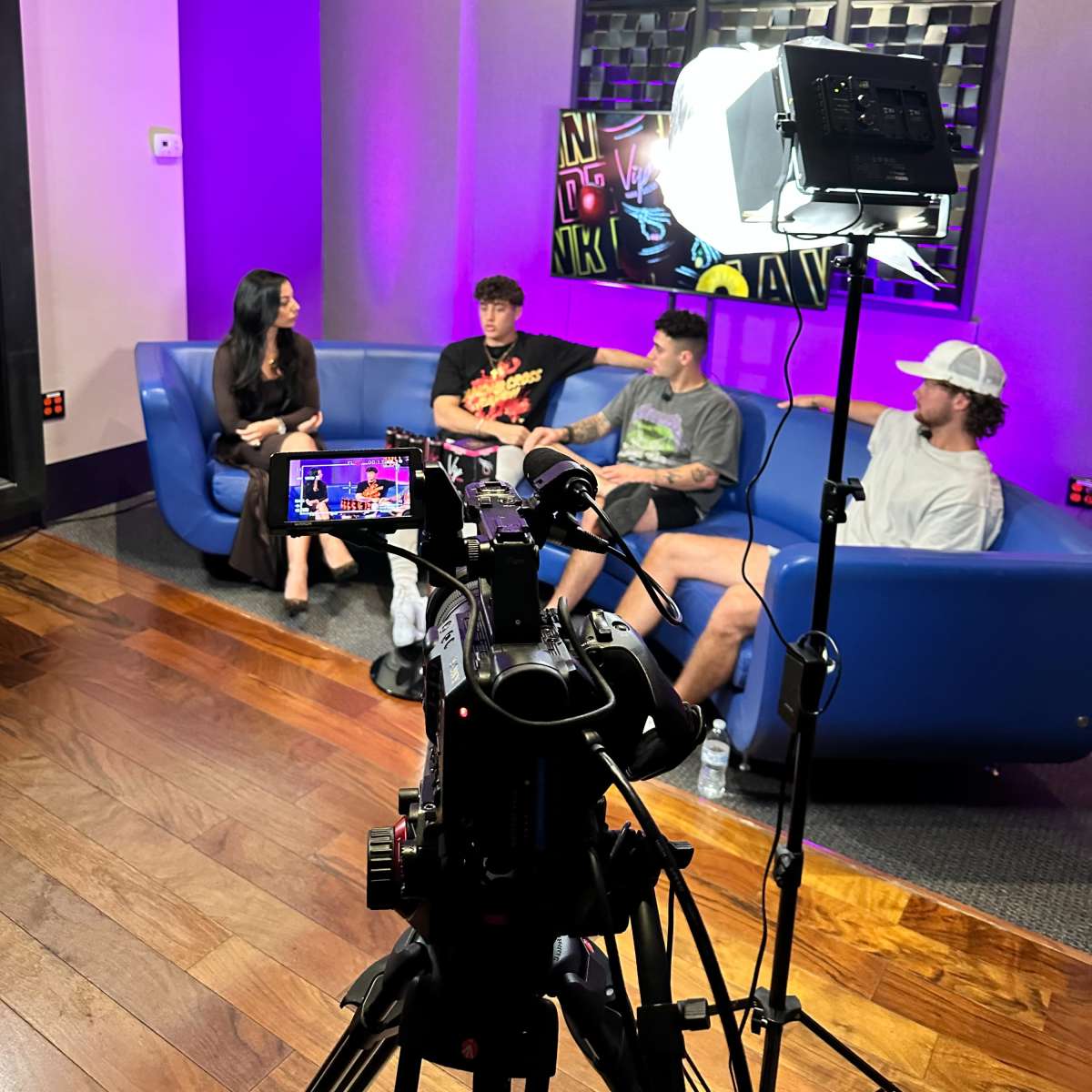A group of four individuals sits on a blue couch in a well-lit room, with a camera set up in front of them for recording.