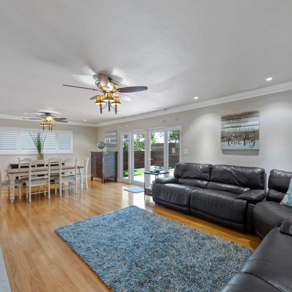 A spacious living room with a large black sectional sofa, a wooden dining table, and access to a backyard through sliding glass doors.
