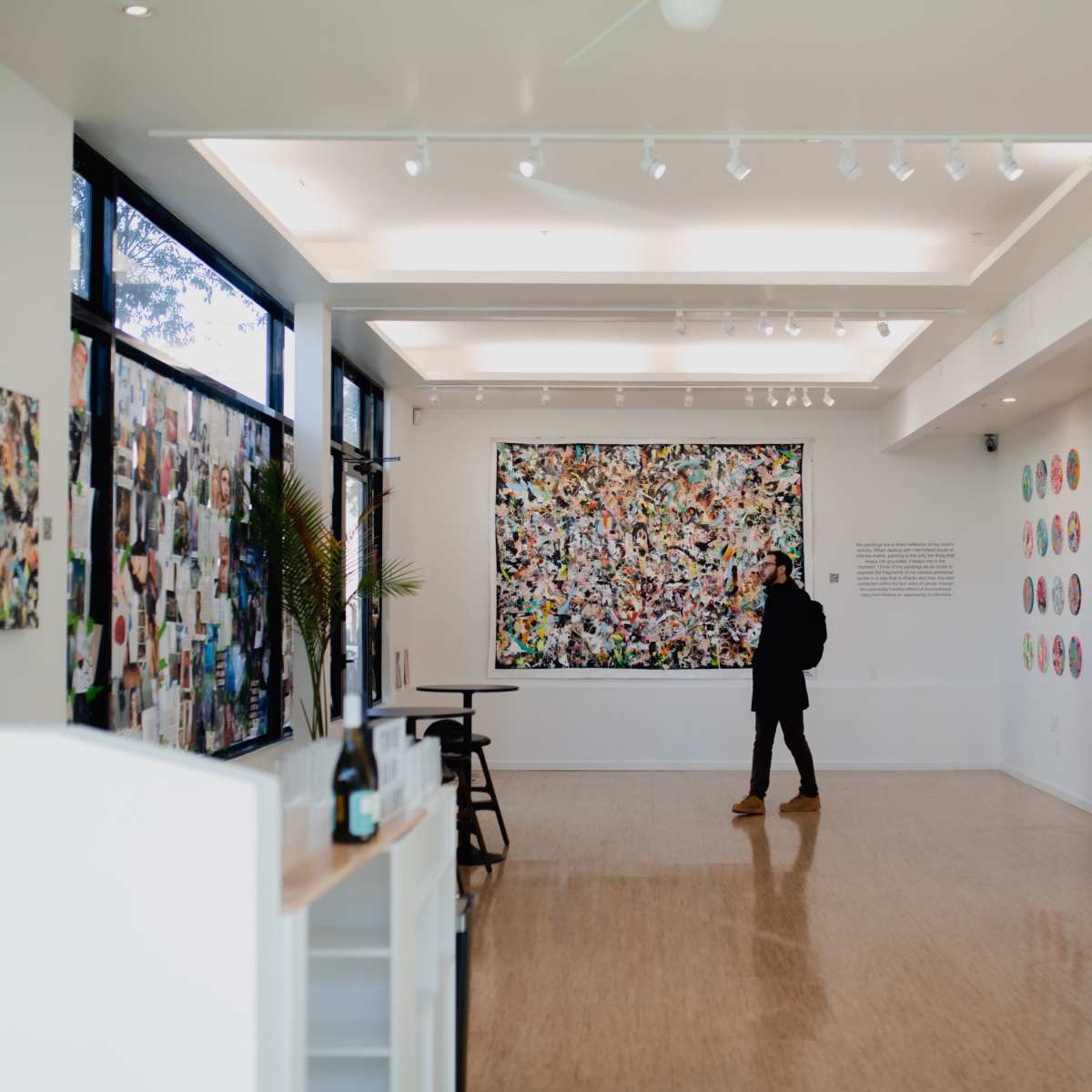 A person stands in an art gallery, observing a large colorful artwork on the wall, surrounded by other art pieces and photographs displayed on the walls.