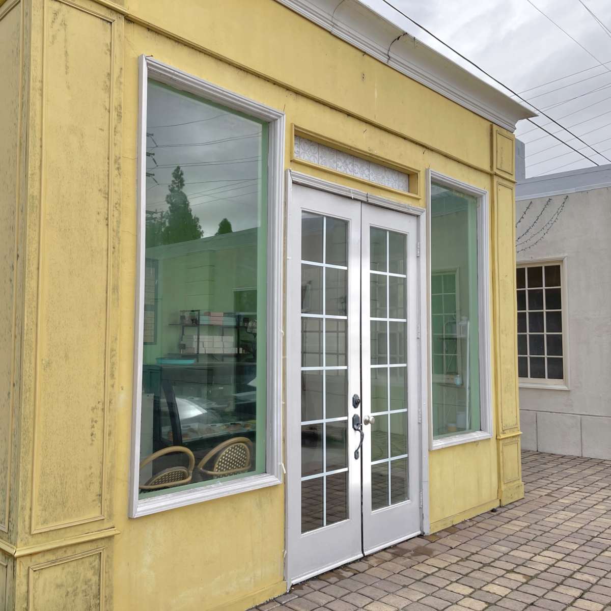A small, yellow building with large glass windows and double doors facing a brick pavement.