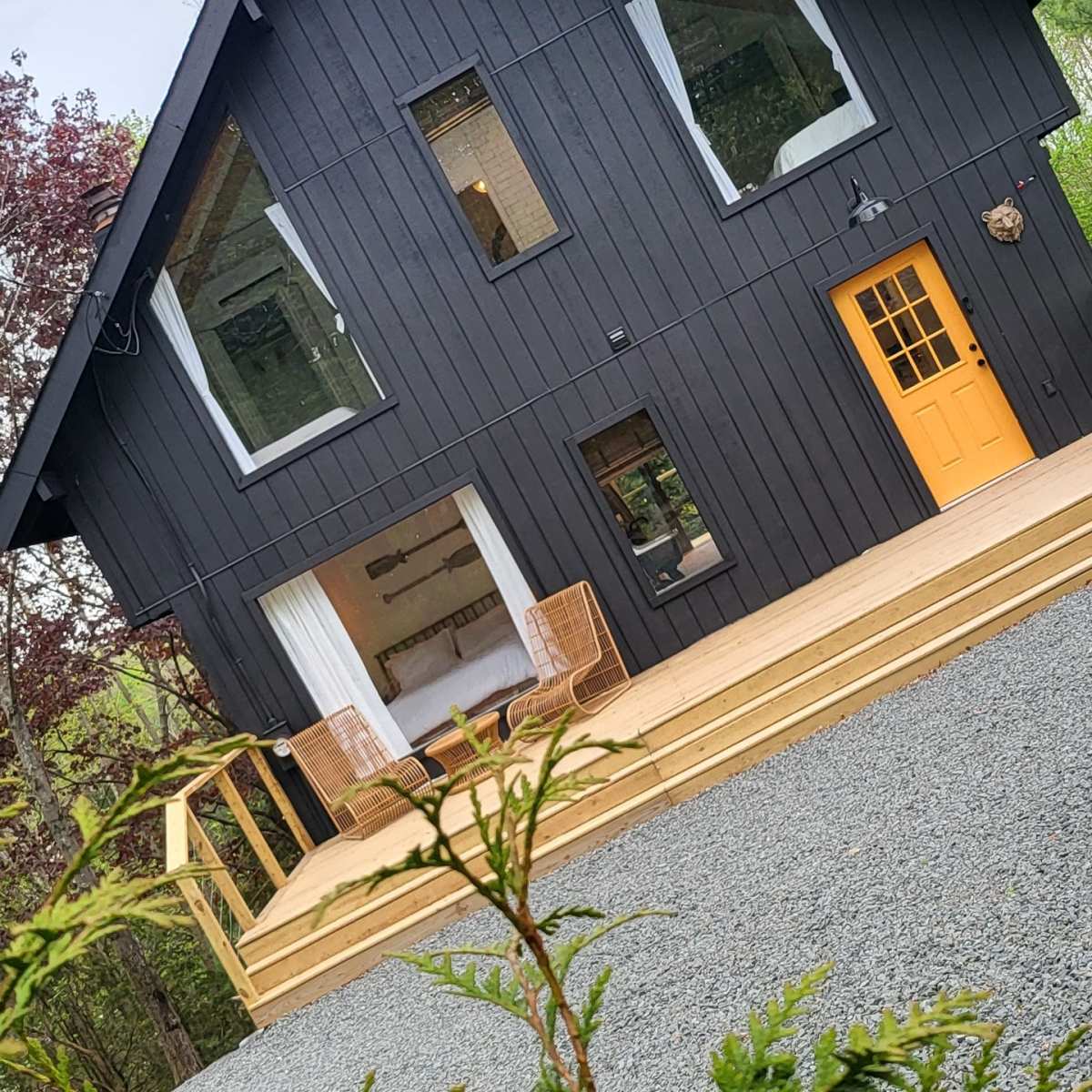 A modern black house with large windows and a wooden deck is situated among trees and a gravel driveway.
