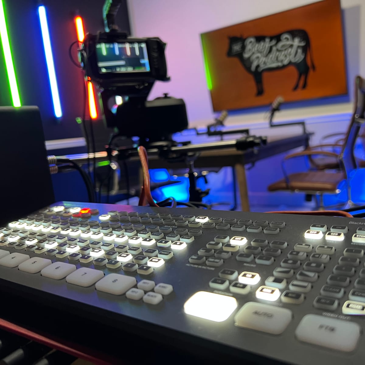 A control panel with illuminated buttons is in the foreground, while a camera and studio setup are visible in the background next to a sign that reads "The Bull & the Tusk."