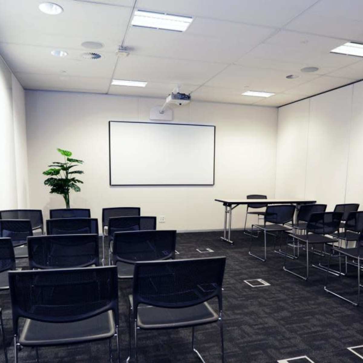 An empty meeting room contains a projector, a blank whiteboard, a table, and rows of black chairs arranged in front.
