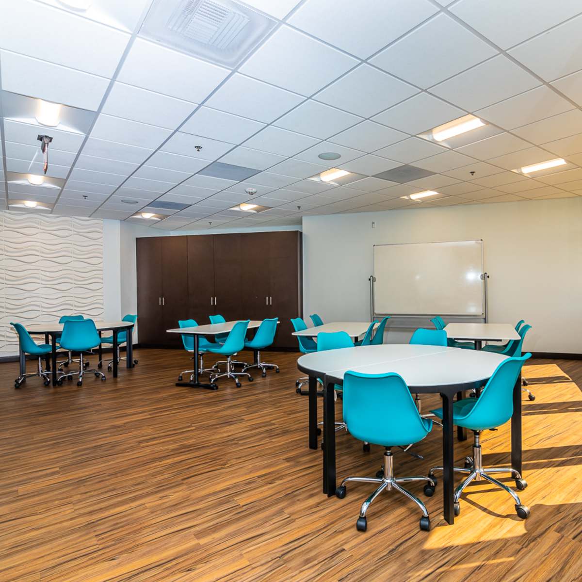 A spacious meeting room with tables and teal chairs, large windows, and a whiteboard.