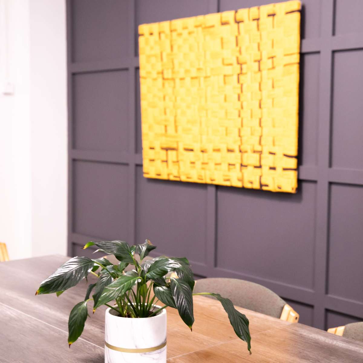 A small potted plant sits on a wooden table in front of a textured yellow wall art piece.