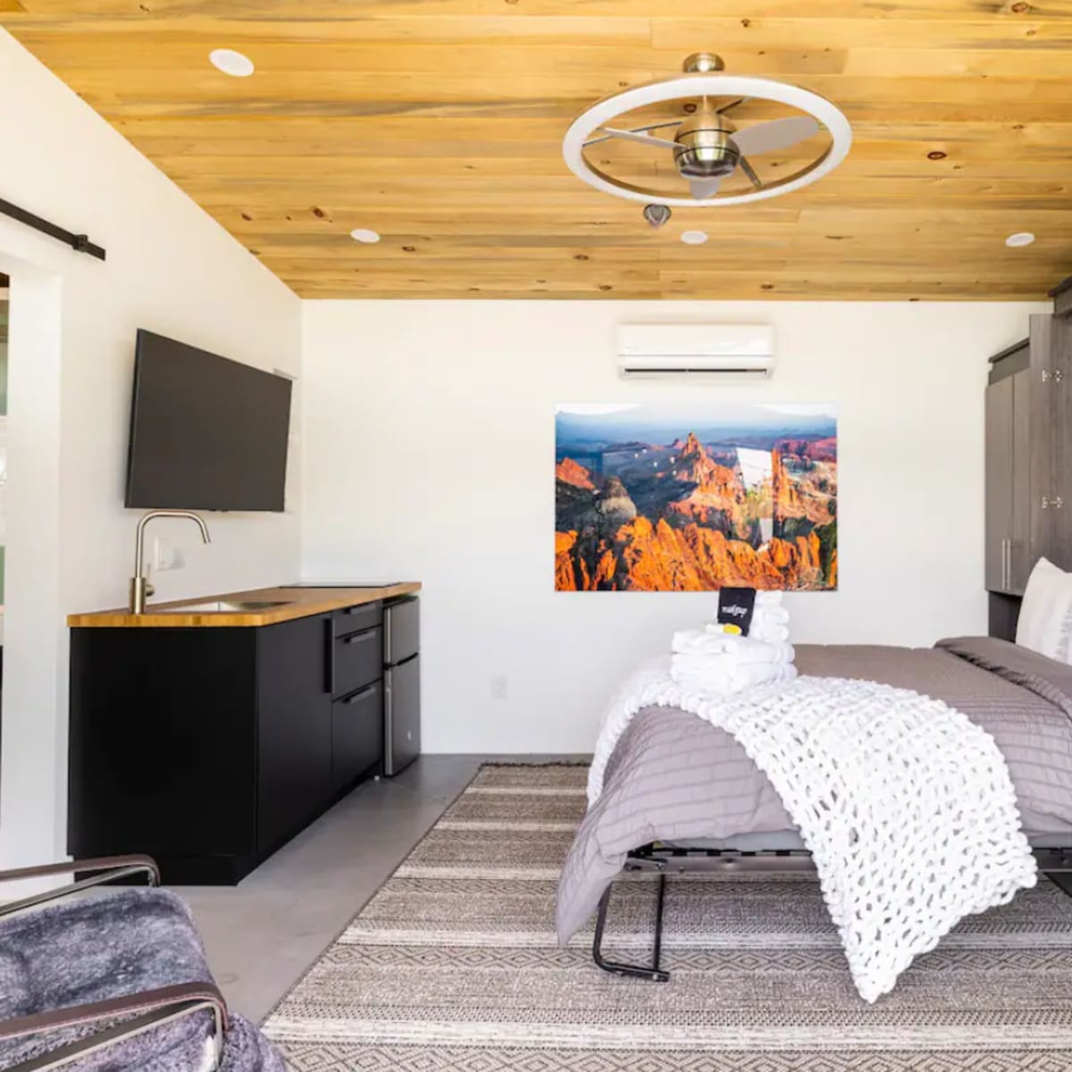A modern, compact bedroom featuring a bed with gray bedding, a wooden ceiling, a flat-screen TV mounted on the wall, and an image of a mountain landscape.