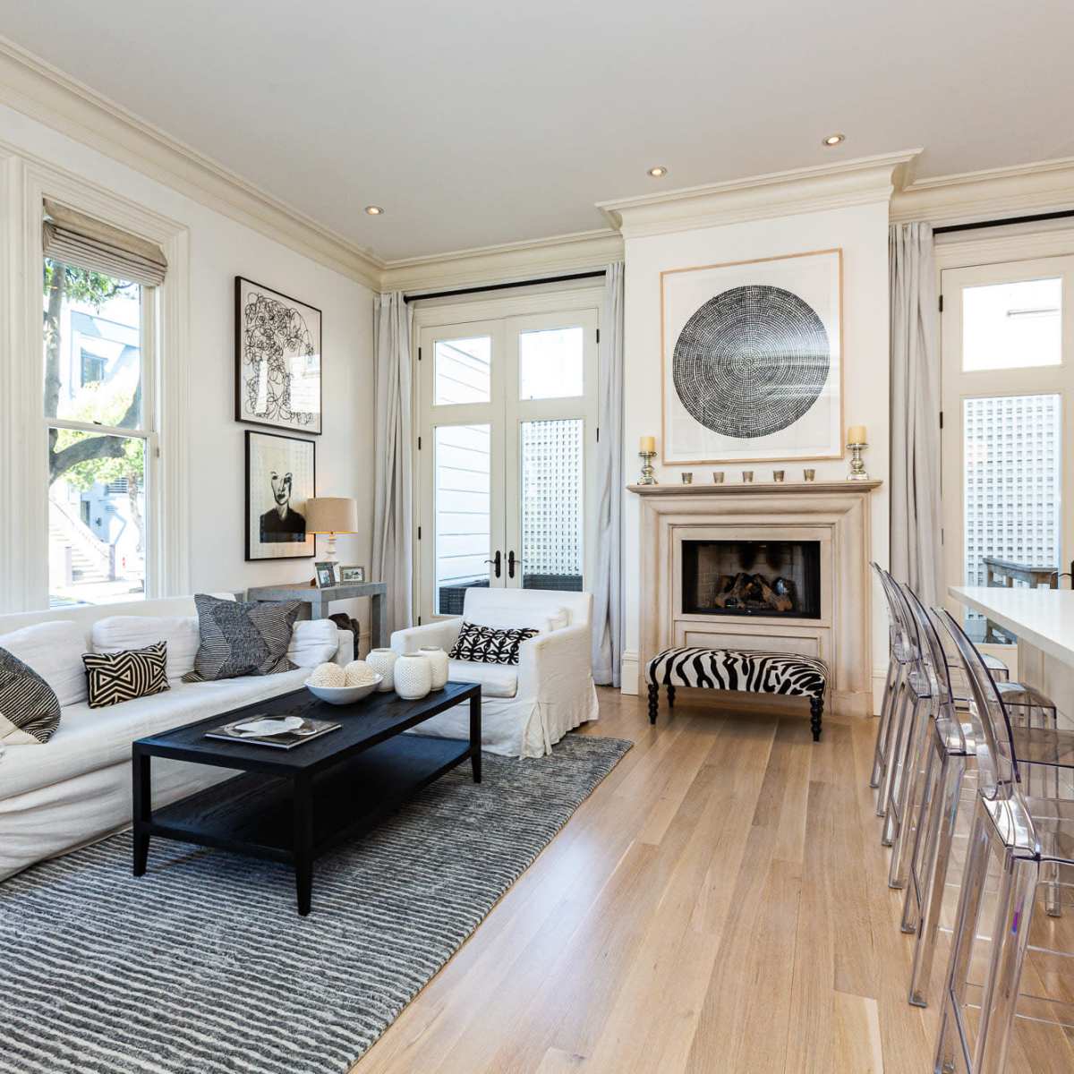 The scene features a light-filled living room with a white sofa, black coffee table, and a fireplace, adjacent to a kitchen bar with clear stools.
