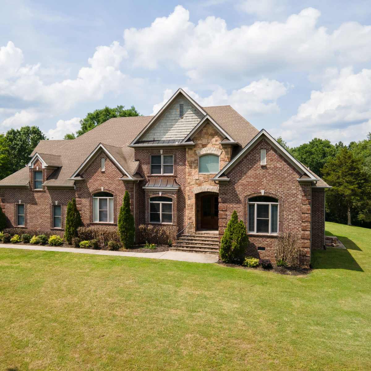 A large brick house with a gabled roof and manicured landscaping sits on a grassy lot under a partly cloudy sky.