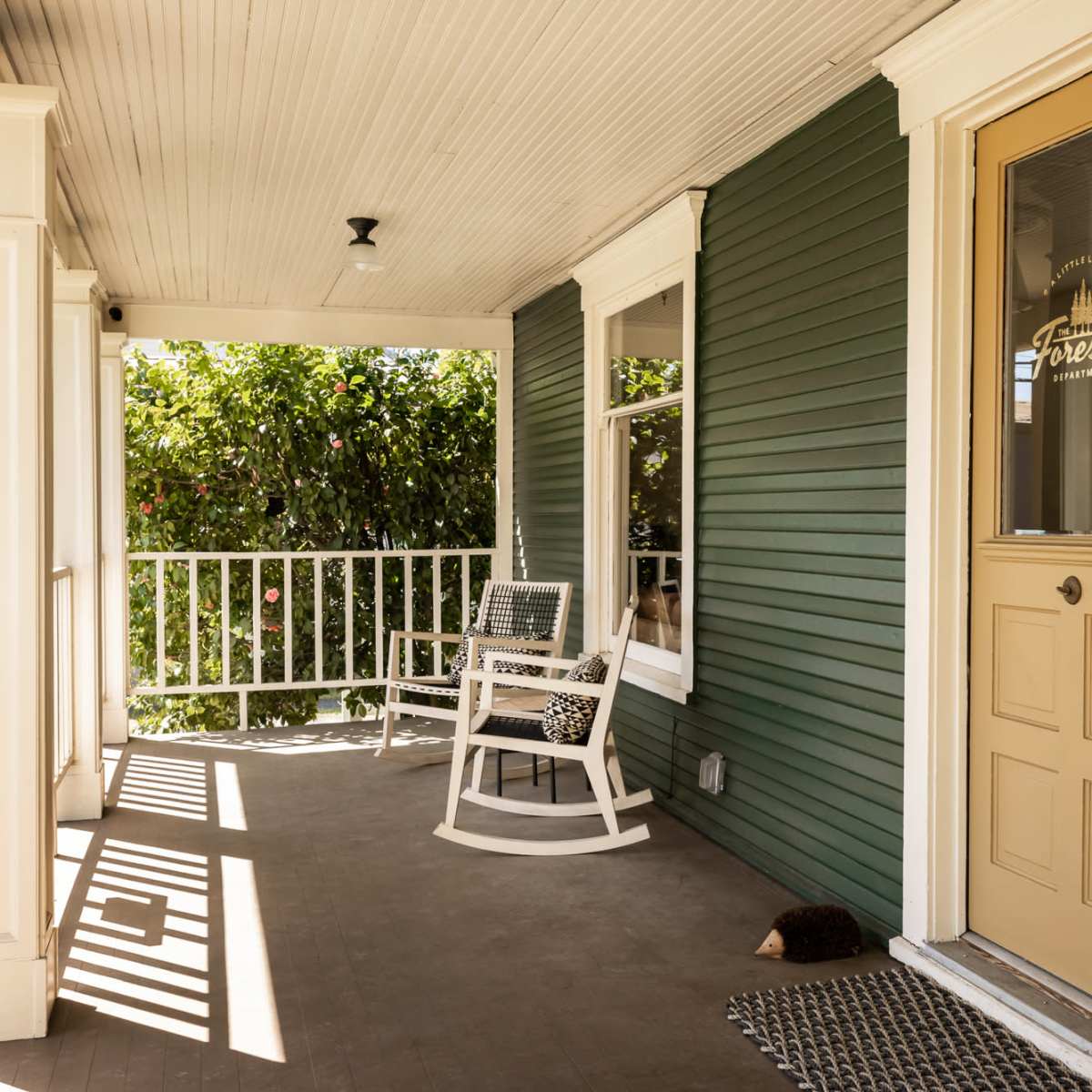 A green porch with a rocking chair, a door numbered 137, and a view of greenery outside.